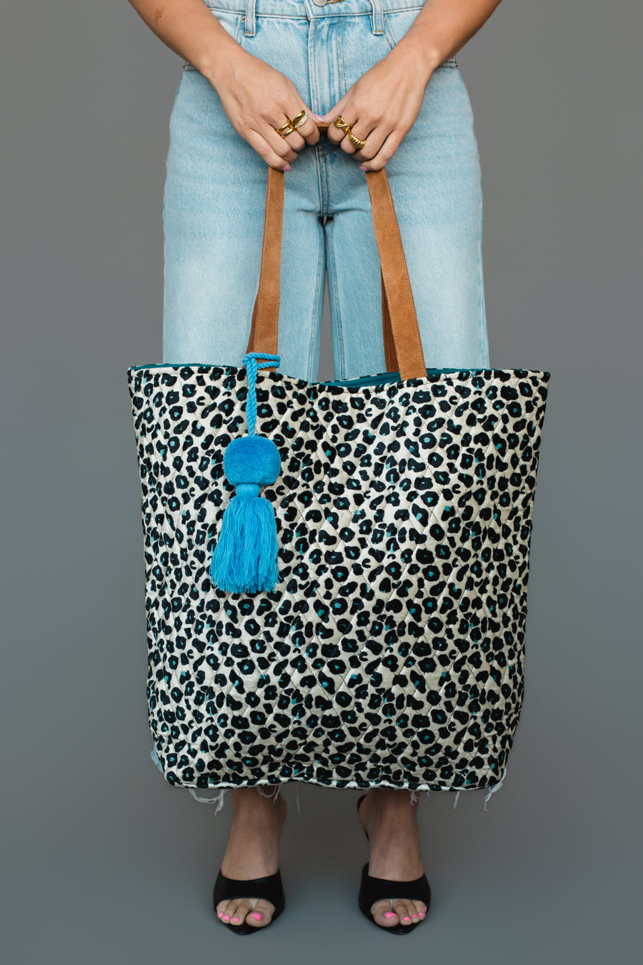 A person in light blue jeans and black sandals holds the Panache Apparel Collection Black, Cream & Blue Leopard Tote, featuring brown handles and a blue tassel pom-pom accessory, against a plain gray background.