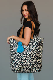 A woman holds the Panache Apparel Collection Black, Cream & Blue Leopard Tote, featuring tan straps and a bright blue tassel, styled with a black top and light blue jeans against a plain background.