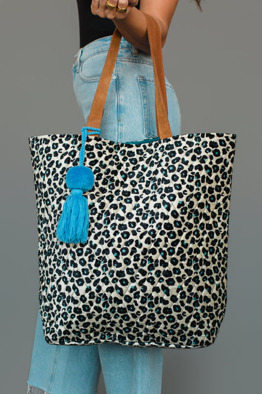 A person holds the Panache Apparel Collection Black, Cream & Blue Leopard Tote, featuring brown handles and a bright blue tassel, styled with light blue jeans and a black top.