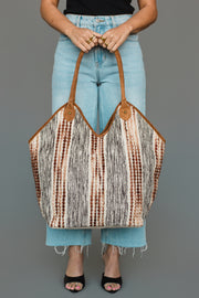A person in light blue jeans and black sandals holds the Panache Apparel Collection Gray & Brown Textured Tote, featuring brown handles and a bold striped, dotted pattern in black, white, and brown hues.