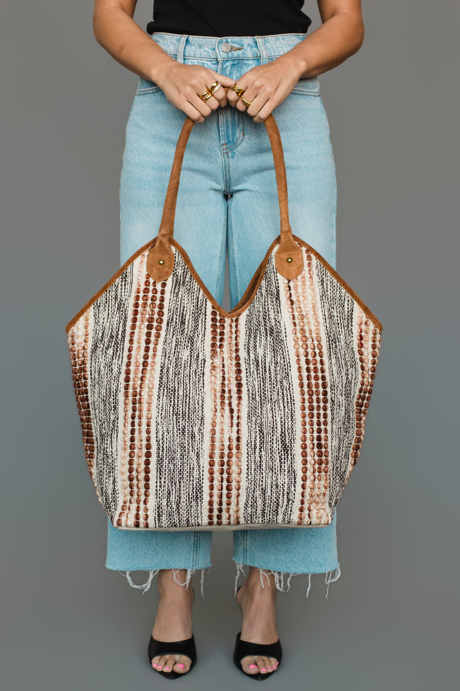 A person in light blue jeans and black sandals holds the Panache Apparel Collection Gray & Brown Textured Tote, featuring brown handles and a bold striped, dotted pattern in black, white, and brown hues.