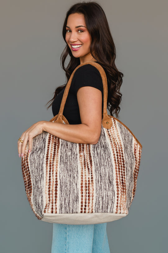 A woman with long brown hair, dressed in a black top and light blue jeans, smiles while carrying the Panache Apparel Collection’s Gray & Brown Textured Tote over her shoulder against a neutral background.