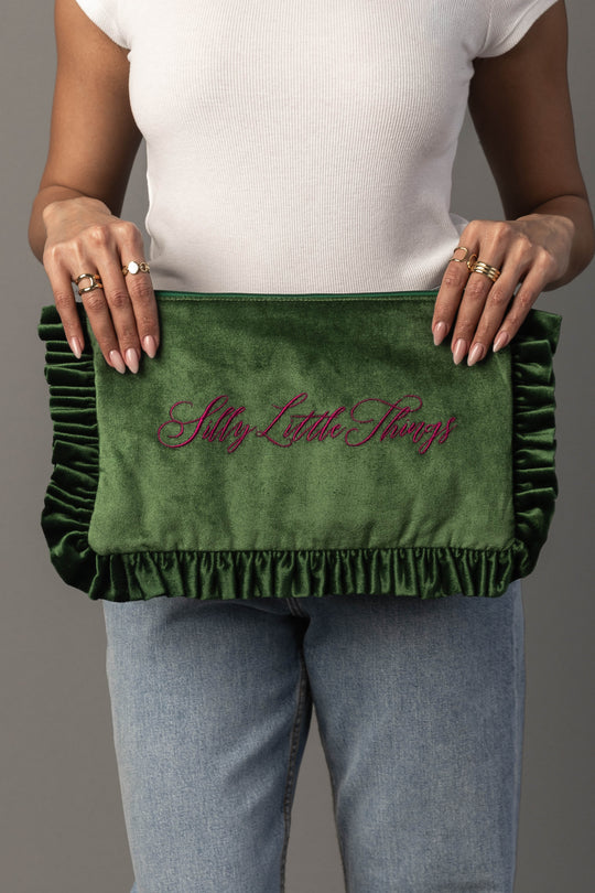 A person wearing a white shirt and blue jeans holds the Panache Apparel Collection’s Silly Little Things Ruffle Travel Pouch, a green velvet clutch with pink script embroidery.