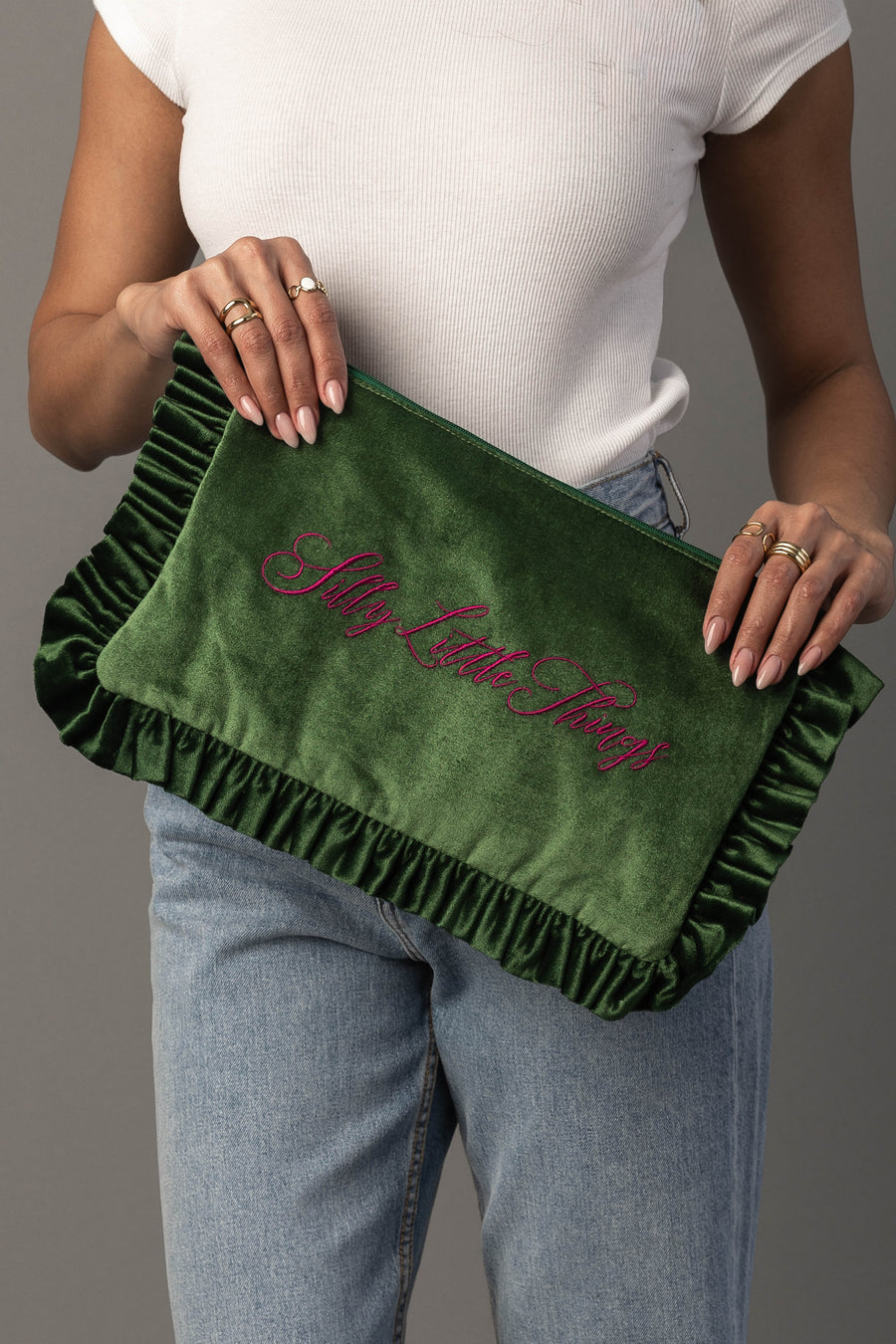 A person in a white shirt and jeans holds the Silly Little Things Ruffle Travel Pouch by Panache Apparel Collection, a green velvet clutch with ruffled edges and “Silly Little Things” embroidered in pink script.