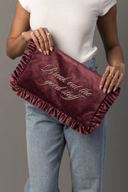 A person in a white top and jeans holds the Panache Apparel Collection’s Break Out The Good Stuff Ruffle Travel Pouch, a maroon velvet clutch with ruffled edges and elegant cursive text that reads, “Break out the good stuff.”.
