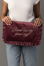 A person in a white shirt and jeans holds the Break Out The Good Stuff Ruffle Travel Pouch from Panache Apparel Collection, a burgundy velvet pouch with ruffled edges and “Break out the good stuff” embroidered in cursive on it.