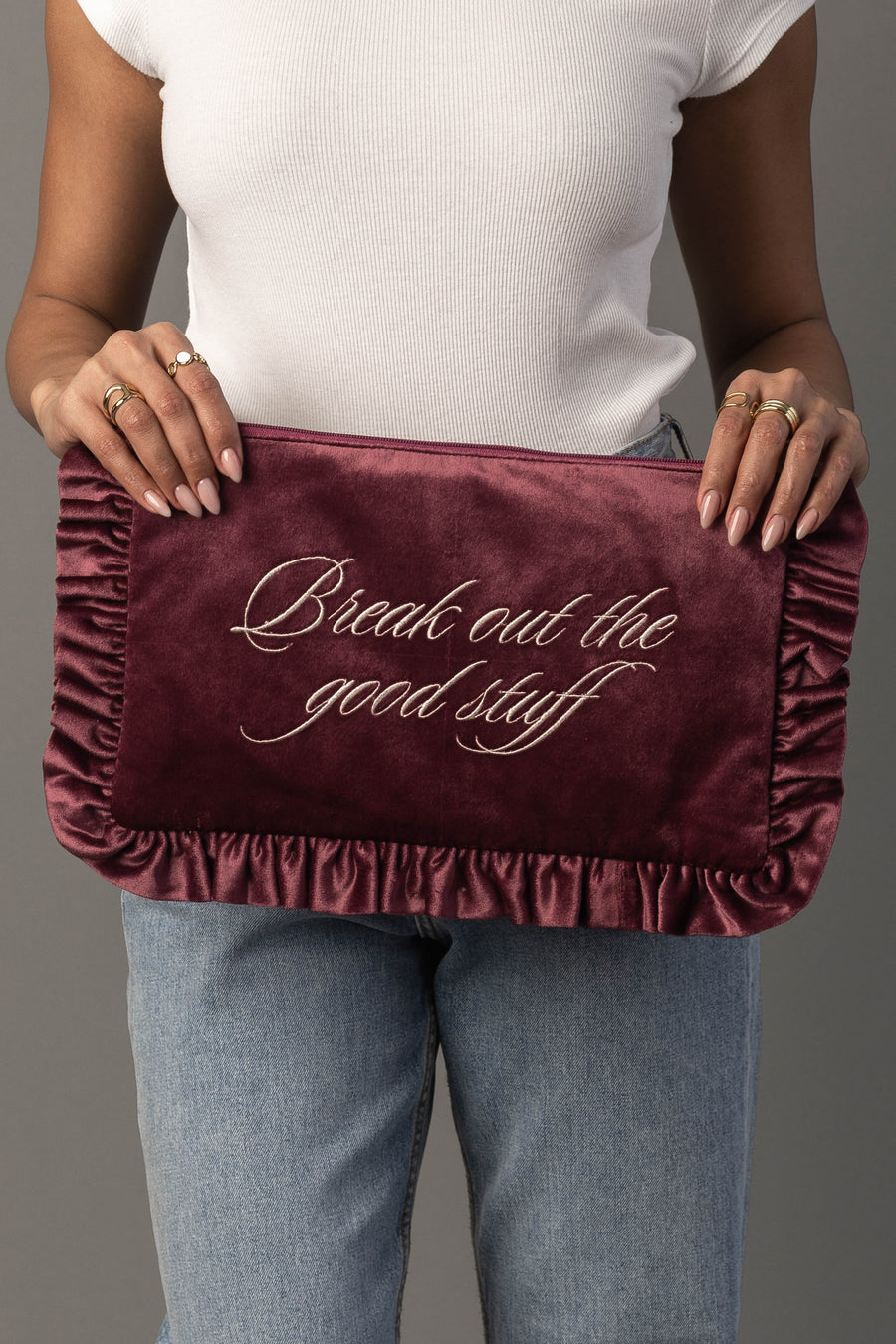 A person in a white shirt and jeans holds the Break Out The Good Stuff Ruffle Travel Pouch from Panache Apparel Collection, a burgundy velvet pouch with ruffled edges and “Break out the good stuff” embroidered in cursive on it.