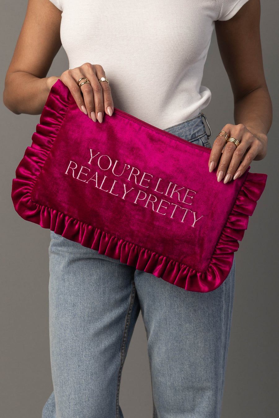 A person in a white sleeveless top and jeans holds the You’re Like Really Pretty Ruffle Travel Pouch by Panache Apparel Collection, a pink velvet clutch with ruffled edges and the words “YOU’RE LIKE REALLY PRETTY” embroidered in white letters.