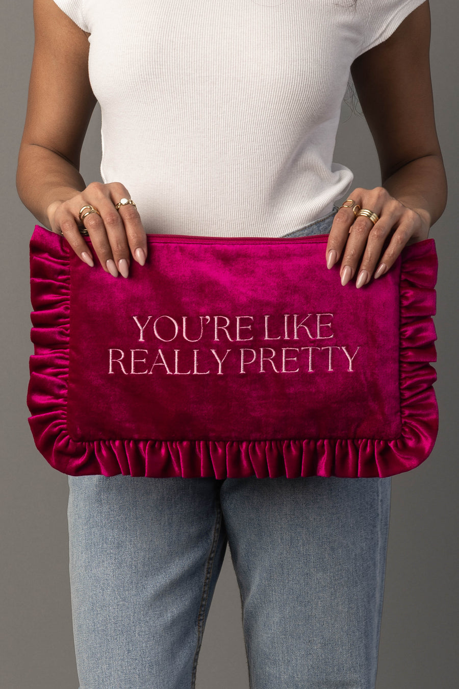A person in a white top and jeans holds a large, ruffled hot pink clutch with the words YOURE LIKE REALLY PRETTY embroidered on it in white letters.