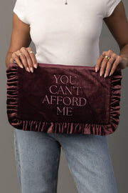 A person in a white shirt and blue jeans holds the Panache Apparel Collection’s You Can’t Afford Me Ruffle Travel Pouch, a large dark red velvet clutch embroidered with the phrase “YOU CANT AFFORD ME” in pink lettering.