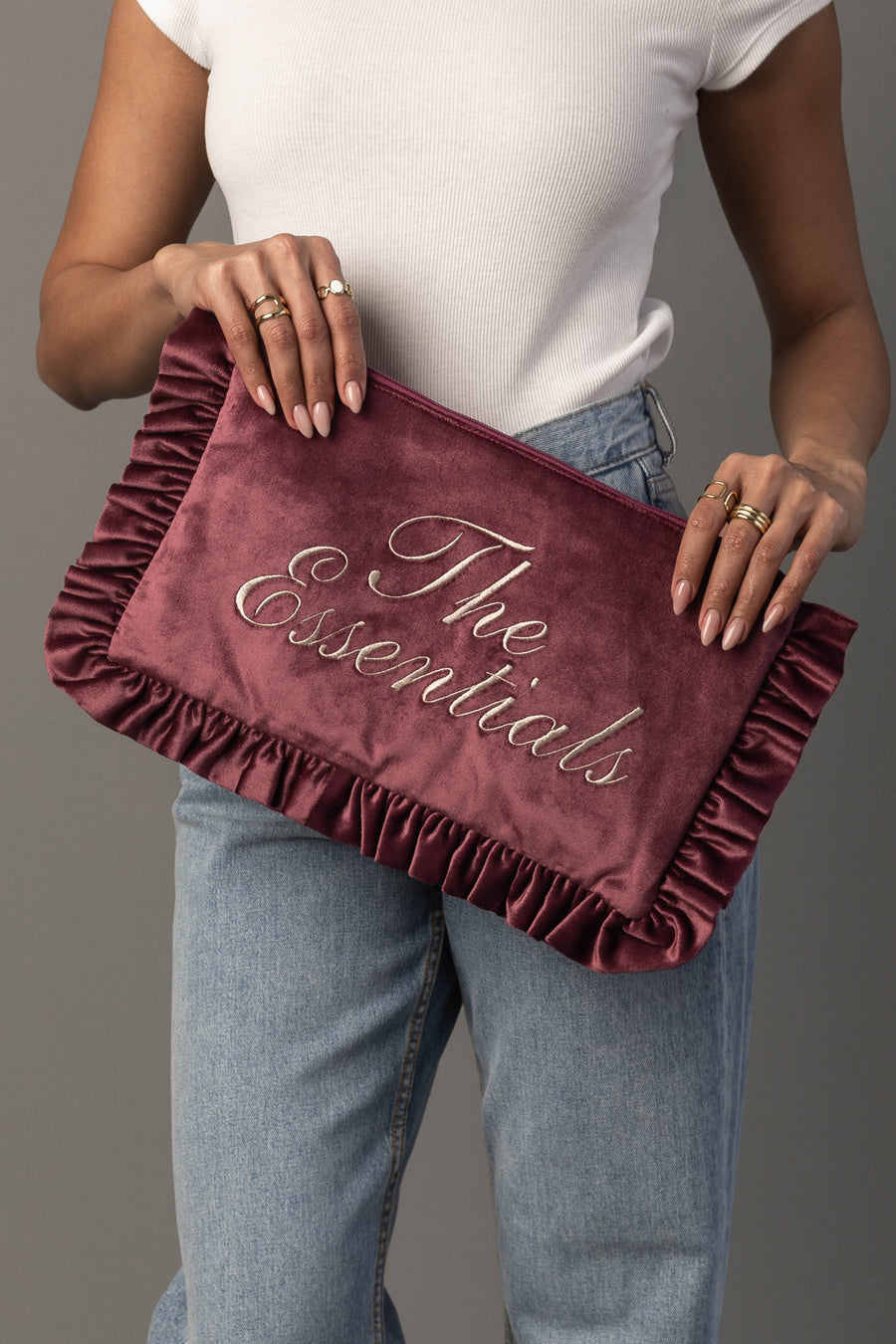 A person wearing a white sleeveless top and jeans holds the Panache Apparel Collection’s The Essentials Ruffle Travel Pouch, a large, burgundy velvet bag with ruffled edges and “The Essentials” written in script.