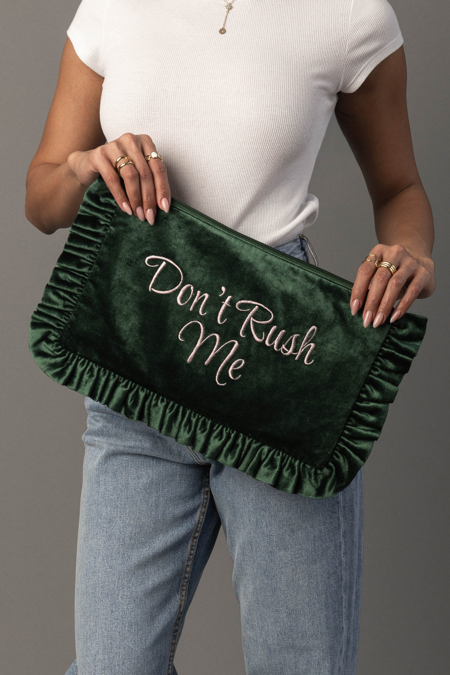 A person in a white shirt and jeans holds the Panache Apparel Collection Don’t Rush Me Ruffle Travel Pouch, a large dark green velvet clutch with ruffled edges and “Don’t Rush Me” embroidered in cursive.