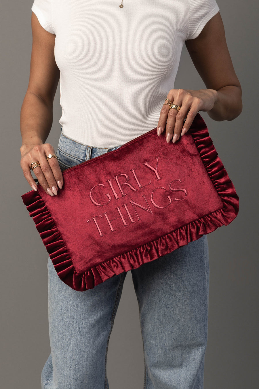 A person in a white shirt and jeans holds the Panache Apparel Collection Girly Things Ruffle Travel Pouch—a red velvet bag with ruffled edges and “GIRLY THINGS” embroidered on it.