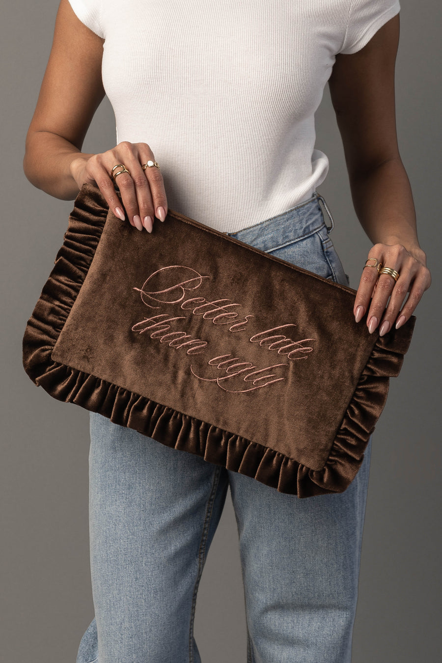 A person wearing a white top and blue jeans holds the Panache Apparel Collection “Better Late Than Ugly Ruffle Travel Pouch,” a brown velvet clutch with ruffled edges and pink script embroidery.