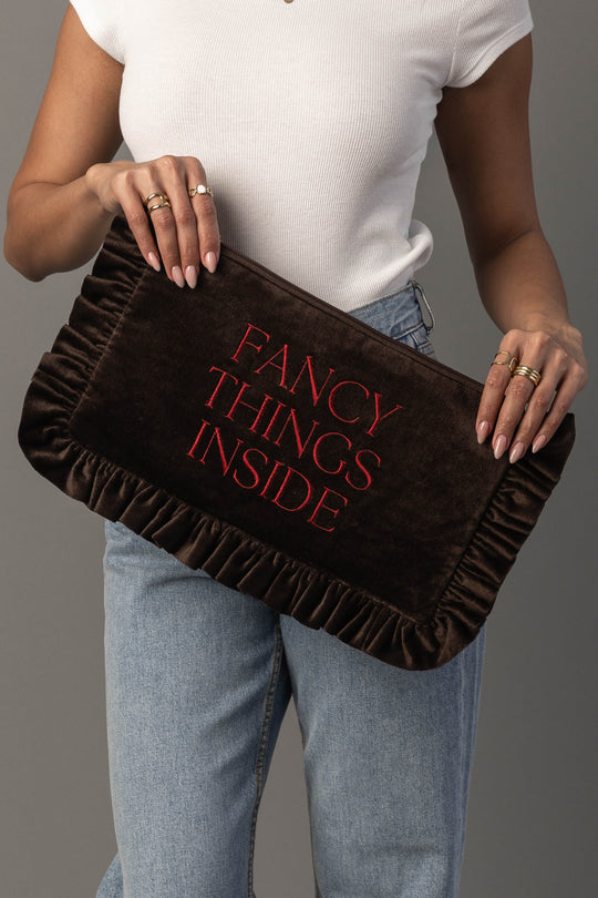 A person in a white top and jeans holds the Panache Apparel Collection’s Fancy Things Inside Ruffle Travel Pouch, a large brown clutch with ruffled edges and “FANCY THINGS INSIDE” written in red letters.