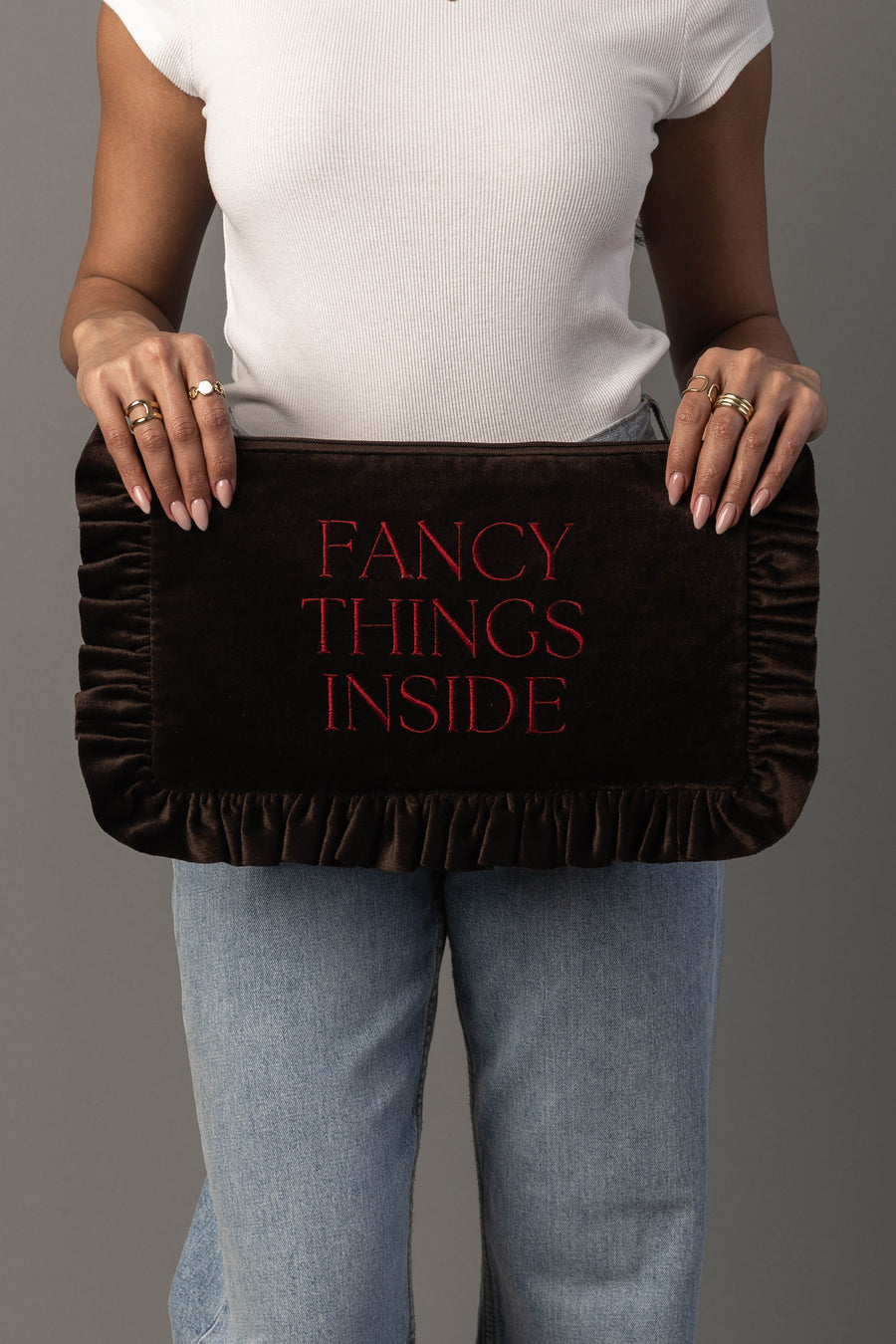 A person in a white shirt and jeans holds the Fancy Things Inside Ruffle Travel Pouch by Panache Apparel Collection, a dark brown velvet pouch with red text that reads “FANCY THINGS INSIDE.”.
