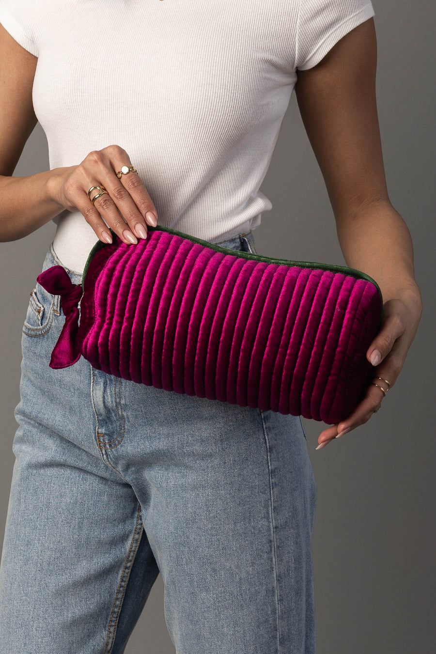 A person in a white shirt and jeans holds the Panache Apparel Collection Fuchsia & Green Ribbed Travel Pouch, which features bright pink ribbed texture, horizontal stitching, a zippered closure, and a velvet ribbon tied to the zipper.