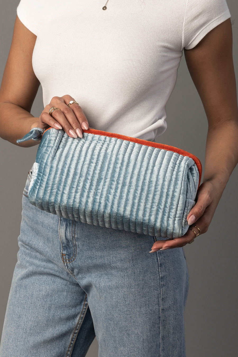 A person in a white shirt and blue jeans holds the Panache Apparel Collection Light Blue & Orange Ribbed Travel Pouch, featuring an orange zipper accent, positioned in front of their waist.