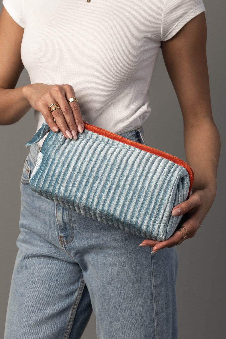 A person in a white ribbed top and blue jeans holds the Panache Apparel Collection’s Light Blue & Orange Ribbed Travel Pouch, featuring a textured design and vibrant orange zipper. Several rings adorn their fingers as they stand against a neutral background.