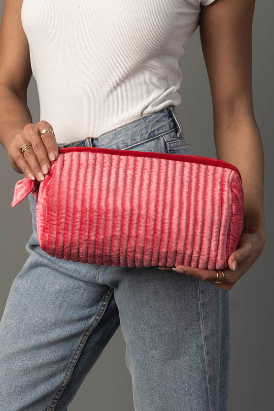 A person in a white shirt and jeans holds the Panache Apparel Collection Pink & Red Ribbed Travel Pouch, which features a rectangular shape, zipper closure, ribbed texture, and a small fabric loop on one end.