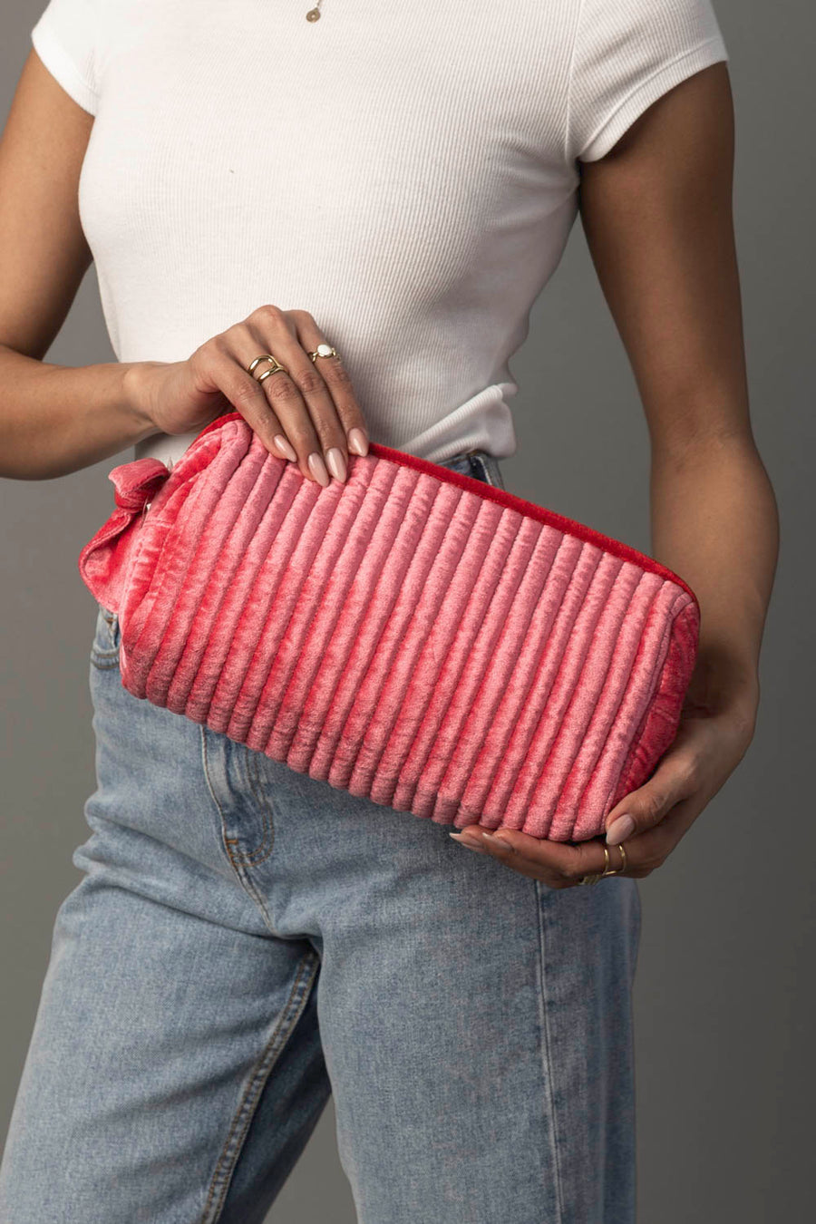 A person in a white short-sleeve top and blue jeans holds the Pink & Red Ribbed Travel Pouch from Panache Apparel Collection with both hands against a plain gray background.