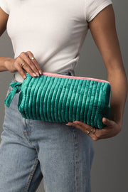 A person dressed in a white shirt and blue jeans holds the Panache Apparel Collection’s Green & Pink Ribbed Travel Pouch, showcasing its quilted green exterior and pink zipper. Multiple rings adorn their fingers, set against a plain gray background.