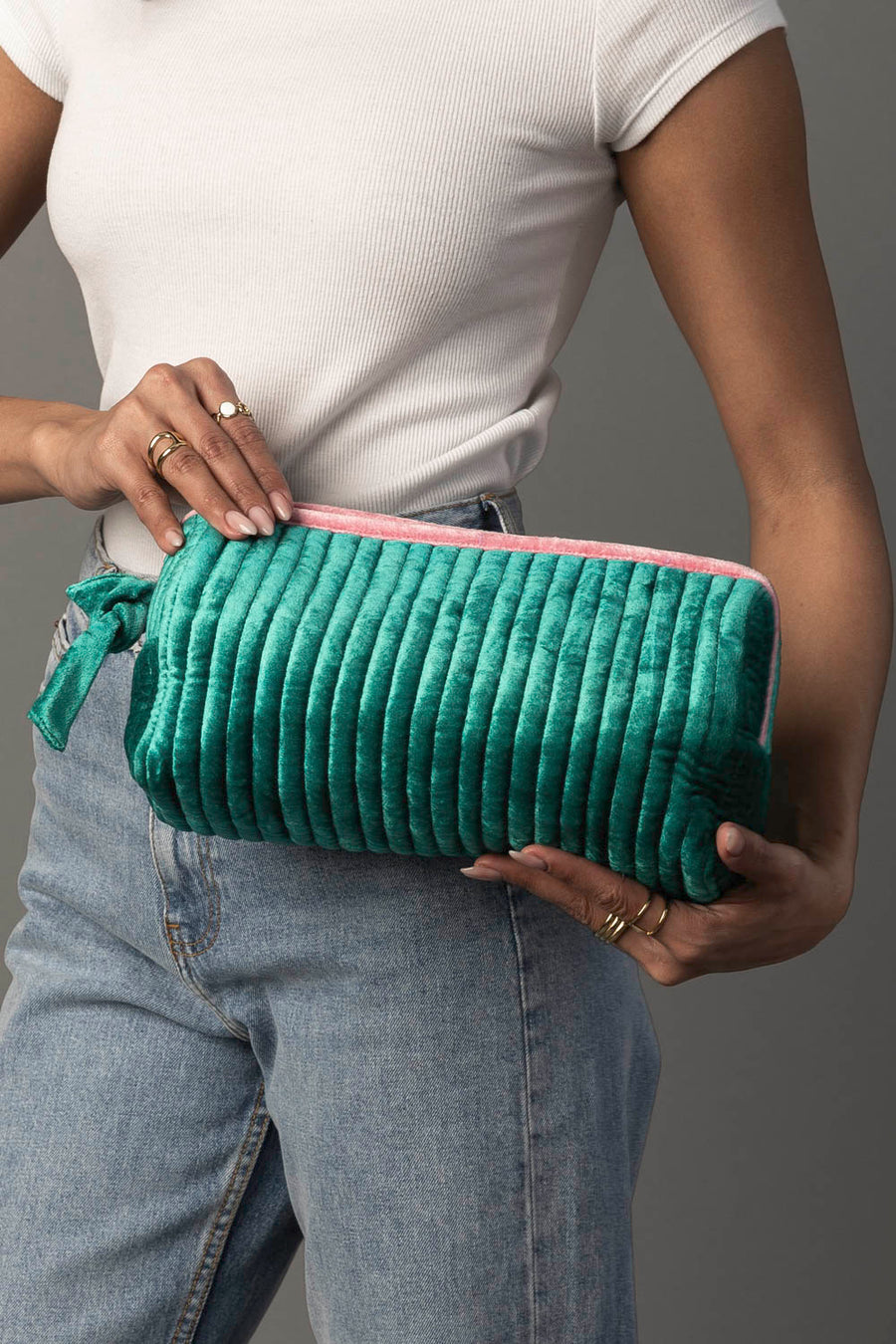 A person dressed in a white shirt and blue jeans holds the Panache Apparel Collection’s Green & Pink Ribbed Travel Pouch, showcasing its quilted green exterior and pink zipper. Multiple rings adorn their fingers, set against a plain gray background.