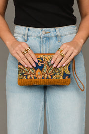 A person wearing light blue jeans and a black top holds the Panache Apparel Collection Mustard & Multicolored Patterned Wristlet, a small bag featuring an intricate design and brown accents. Multiple gold rings adorn their fingers.