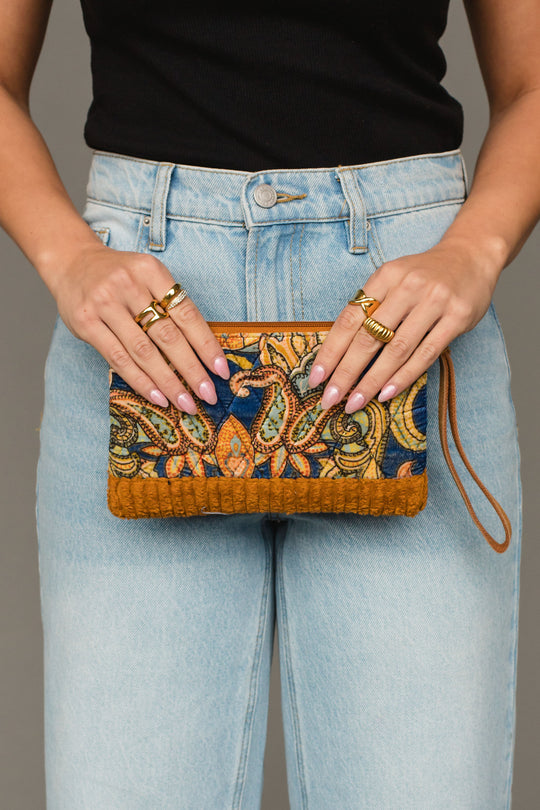 A person wearing light blue jeans and a black top holds the Panache Apparel Collection Mustard & Multicolored Patterned Wristlet, a small bag featuring an intricate design and brown accents. Multiple gold rings adorn their fingers.
