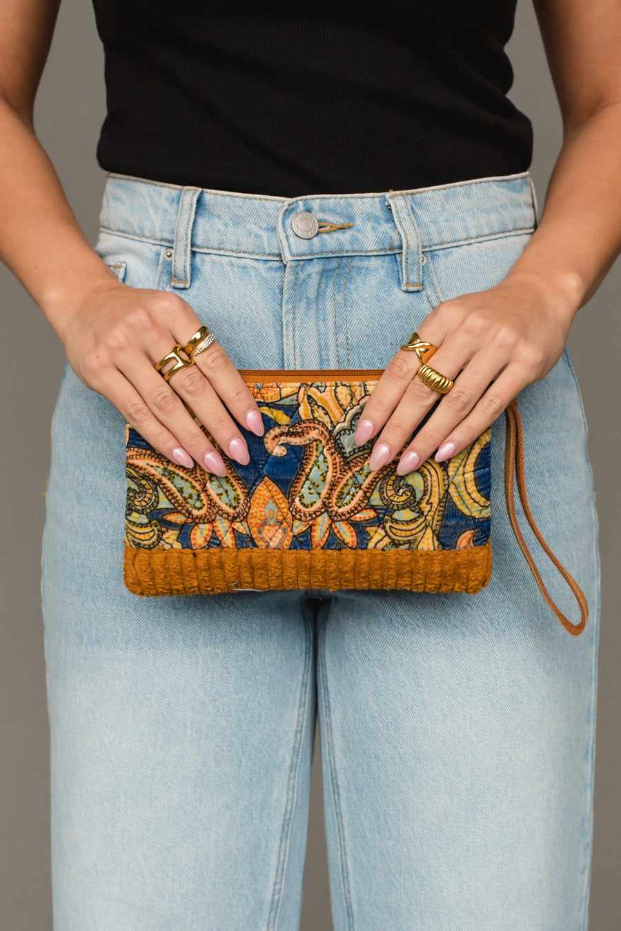 A person wearing light blue jeans and a black top holds the Panache Apparel Collection Mustard & Multicolored Patterned Wristlet, a small bag featuring an intricate design and brown accents. Multiple gold rings adorn their fingers.
