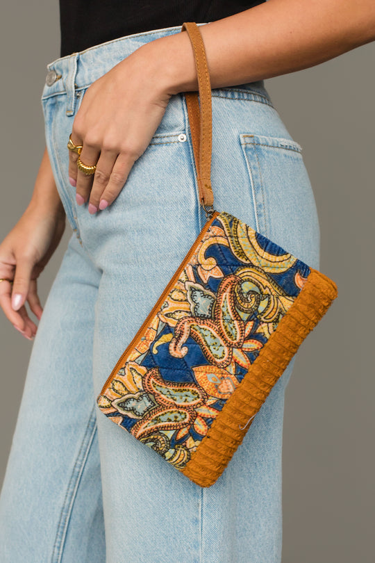 A person in light blue jeans holds the Mustard & Multicolored Patterned Wristlet by Panache Apparel Collection, featuring a brown leather strap and trim.