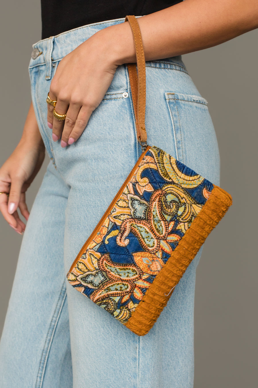 A person in light blue jeans holds the Mustard & Multicolored Patterned Wristlet by Panache Apparel Collection, featuring a brown leather strap and trim.