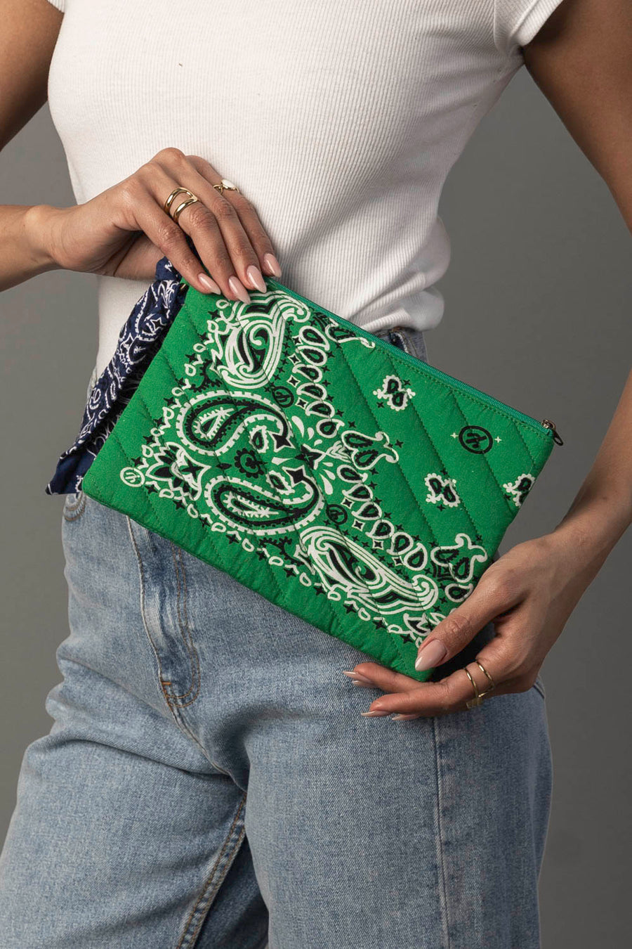 A person in a white tank top and blue jeans holds the Panache Apparel Collection Green Bandana Clutch, which features a blue bandana detail, while displaying several rings on their fingers.