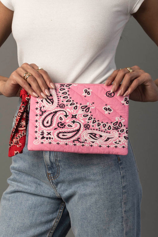 A person wearing a white shirt and blue jeans holds the Pink Bandana Clutch from Panache Apparel Collection. They have several rings on their fingers and a red bandana tied to their belt loop.