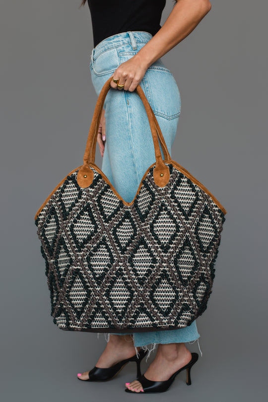 A woman in a black top and light jeans holds the Panache Apparel Collection Black & Gray Diamond Pattern Textured Tote, featuring brown handles. She pairs the look with black heeled sandals.