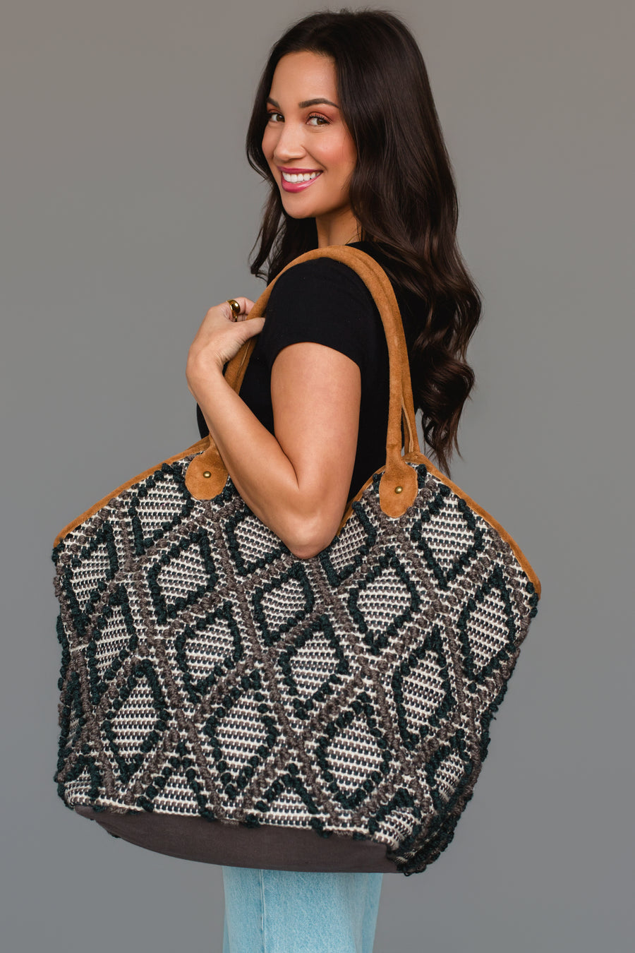 A woman with long dark hair, dressed in a black top and jeans, smiles while holding the Panache Apparel Collection Black & Gray Diamond Pattern Textured Tote, featuring geometric designs and brown handles, against a plain gray background.