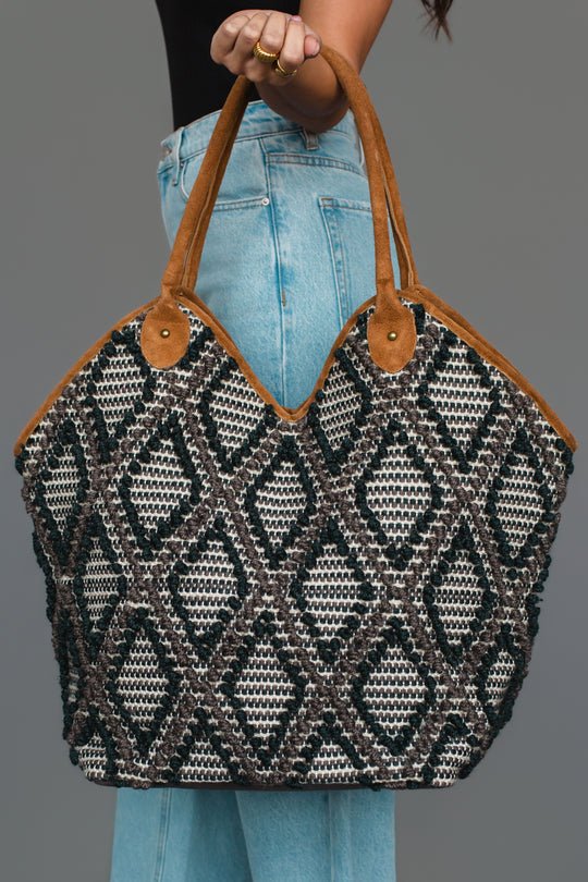 A person wearing light blue jeans holds the Panache Apparel Collection Black & Gray Diamond Pattern Textured Tote, featuring brown suede handles, against a plain gray background.