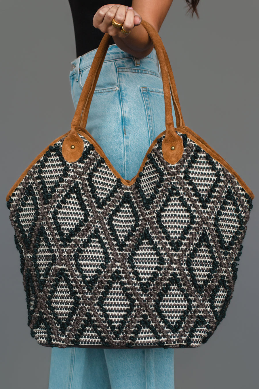 A person wearing light blue jeans holds the Panache Apparel Collection Black & Gray Diamond Pattern Textured Tote, featuring brown suede handles, against a plain gray background.