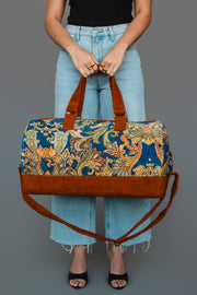 A person in light blue jeans and black sandals holds the Panache Apparel Collection Rust & Multicolored Patterned Duffel, featuring brown handles and strap, against a plain gray background.