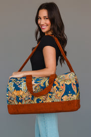 A woman smiles while holding the Panache Apparel Collection Rust & Multicolored Patterned Duffel, featuring bright orange trim and handles. She wears a black top with light blue jeans and stands against a plain gray background.