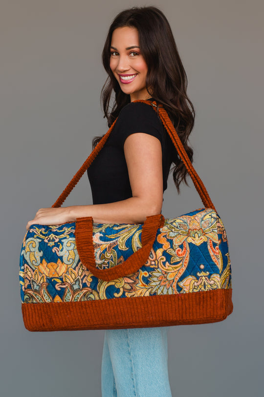 A woman smiles while holding the Panache Apparel Collection Rust & Multicolored Patterned Duffel, featuring bright orange trim and handles. She wears a black top with light blue jeans and stands against a plain gray background.