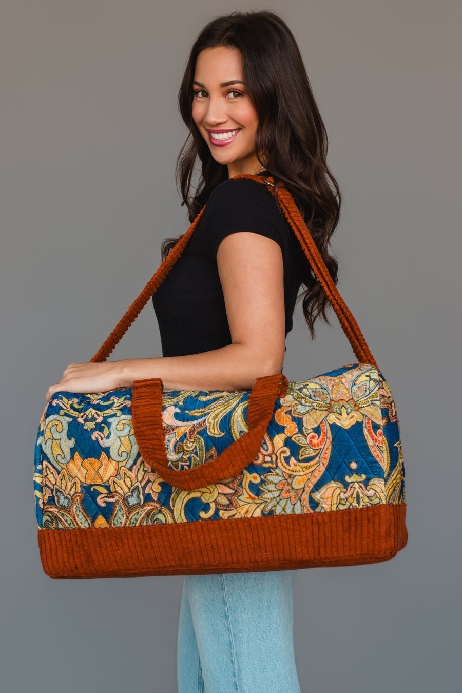 A woman smiles while holding the Panache Apparel Collection Rust & Multicolored Patterned Duffel, featuring bright orange trim and handles. She wears a black top with light blue jeans and stands against a plain gray background.