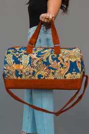 A person in jeans holds the Panache Apparel Collection’s Rust & Multicolored Patterned Duffel, featuring vibrant floral designs and orange handles and trim, against a plain gray background.