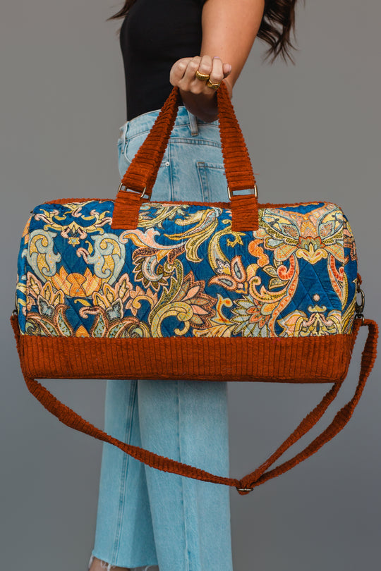 A person in jeans holds the Panache Apparel Collection’s Rust & Multicolored Patterned Duffel, featuring vibrant floral designs and orange handles and trim, against a plain gray background.