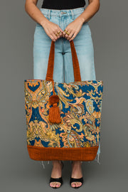 A person in jeans and black sandals holds the Panache Apparel Collection Rust & Multicolored Patterned Tote, featuring vibrant paisley designs, orange accents, and a tassel, against a plain gray background.