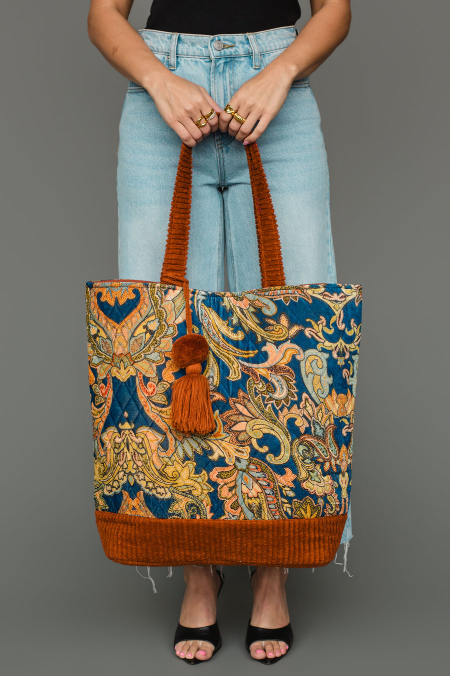 A person in jeans and black sandals holds the Panache Apparel Collection Rust & Multicolored Patterned Tote, featuring vibrant paisley designs, orange accents, and a tassel, against a plain gray background.