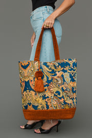 A person in blue jeans and black heels holds the Panache Apparel Collection Rust & Multicolored Patterned Tote, featuring orange handles and a tassel, against a gray background.