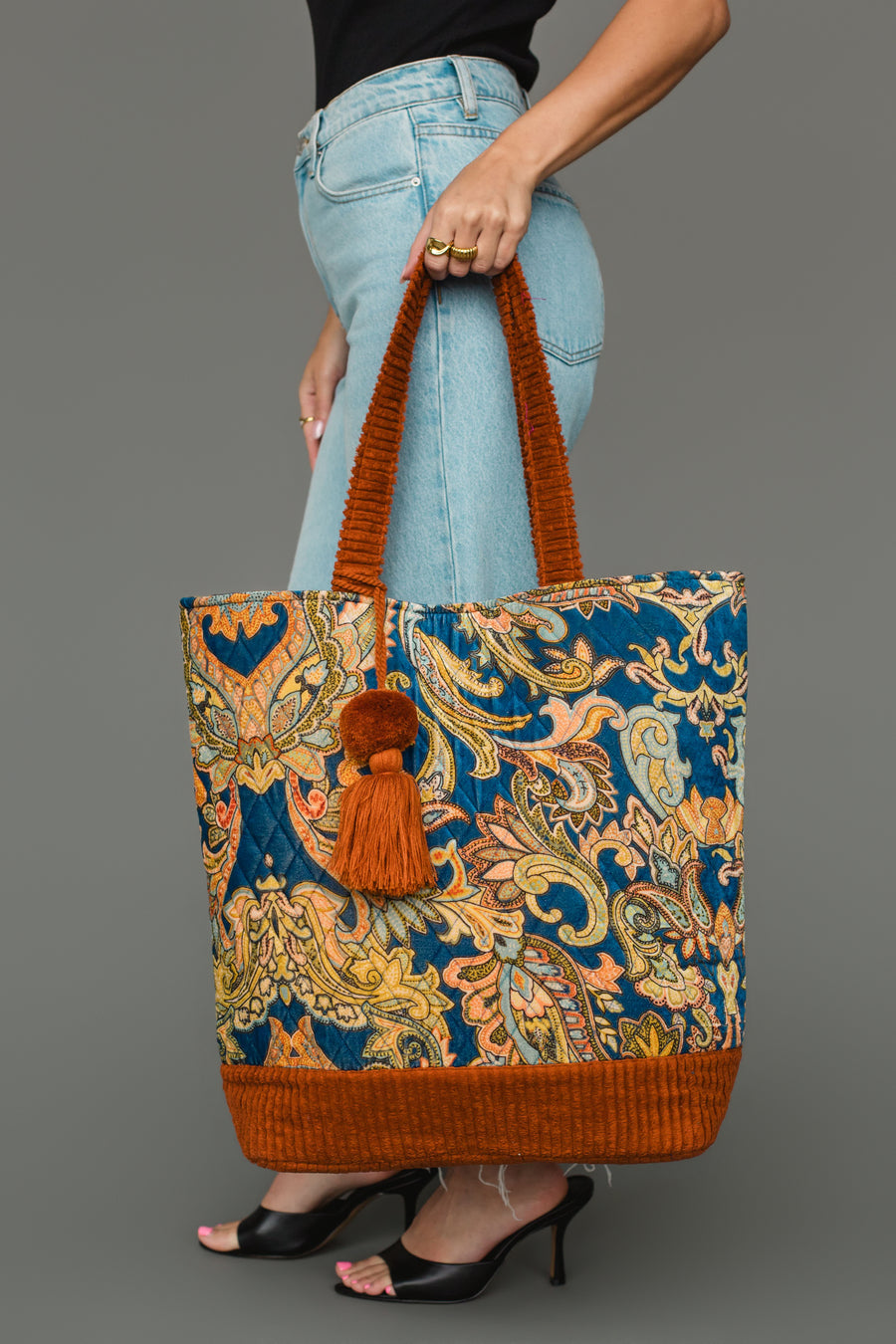 A person in blue jeans and black heels holds the Panache Apparel Collection Rust & Multicolored Patterned Tote, featuring orange handles and a tassel, against a gray background.