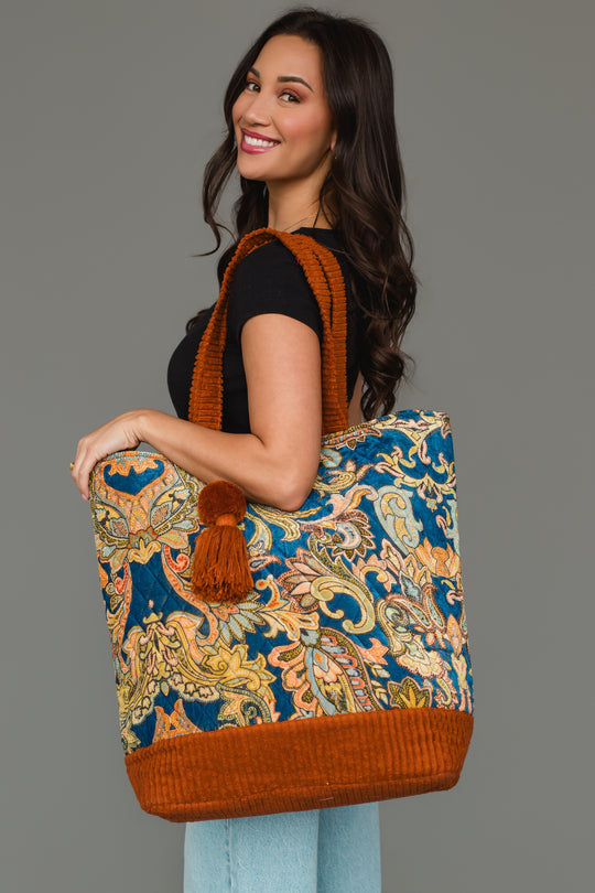 A woman smiles while wearing a black top and light jeans, holding the Panache Apparel Collection’s Rust & Multicolored Patterned Tote—a large paisley-print bag with orange accents and a pom-pom—standing against a plain gray background.