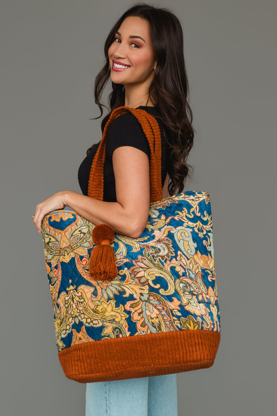 A woman smiles while wearing a black top and light jeans, holding the Panache Apparel Collection’s Rust & Multicolored Patterned Tote—a large paisley-print bag with orange accents and a pom-pom—standing against a plain gray background.